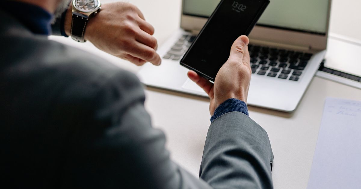 Profissional a verificar as horas no telemóvel e no relógio, com o portátil no escritório